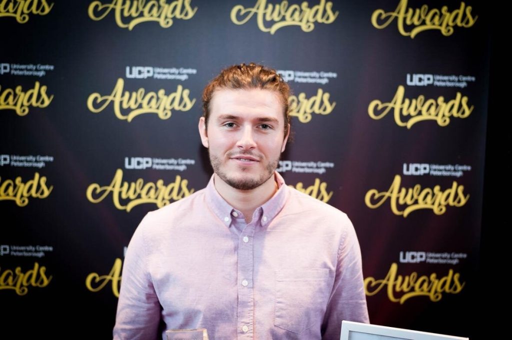 Gabriel Manthorp in a light pink/red shirt holding an award with a Peterborough College backdrop behind him. He's looking at the camera with a gentle smile. His brown hair is tied up on his head. 